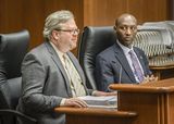David Anderson sits at the testifiers' table and speaks to someone out of view. Representative Noor sits beside him and watches him speak.